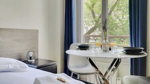 A hotel room with a bed, a dining table with chairs, a vase with flowers, and a view of trees through the window. - Peaceful Studio -2P- Gare de l'Est/République (Paris)