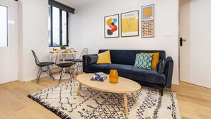 Una sala de estar moderna con un sofá azul oscuro, una mesa de centro redonda de madera y un tapete estampado. Se observan obras de arte abstracto en la pared y un comedor con mesa y sillas.