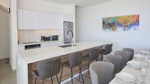 A modern kitchen with white cabinetry, a stainless steel refrigerator, and a bar area with grey chairs. - Protaras Aqua Pearl Villa Aq11 (Paralimni)