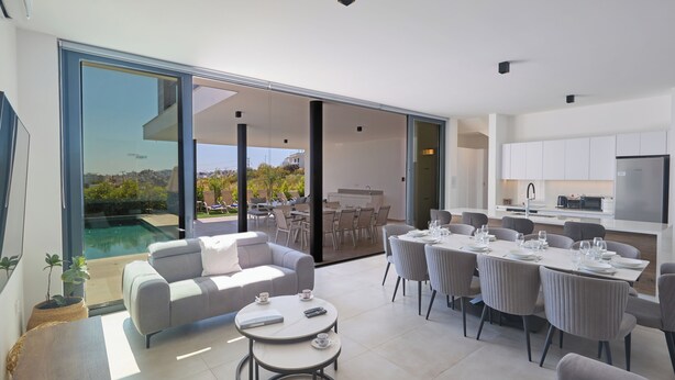 A modern open-plan living area with a dining space, a sofa, and a table with remote controls. - Protaras Aqua Pearl Villa Aq11 (Paralimni)