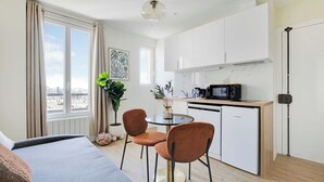 Een compacte hotelkamer met een bed, een klein rond tafeltje met twee stoelen, een kitchenette en een groot raam met gordijnen.