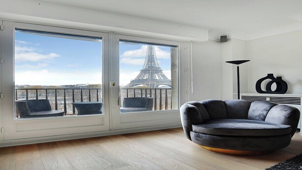 Un saló modern amb vistes a la Torre Eiffel a través de grans finestres, un sofà de cuir negre i una làmpada de peu minimalista.