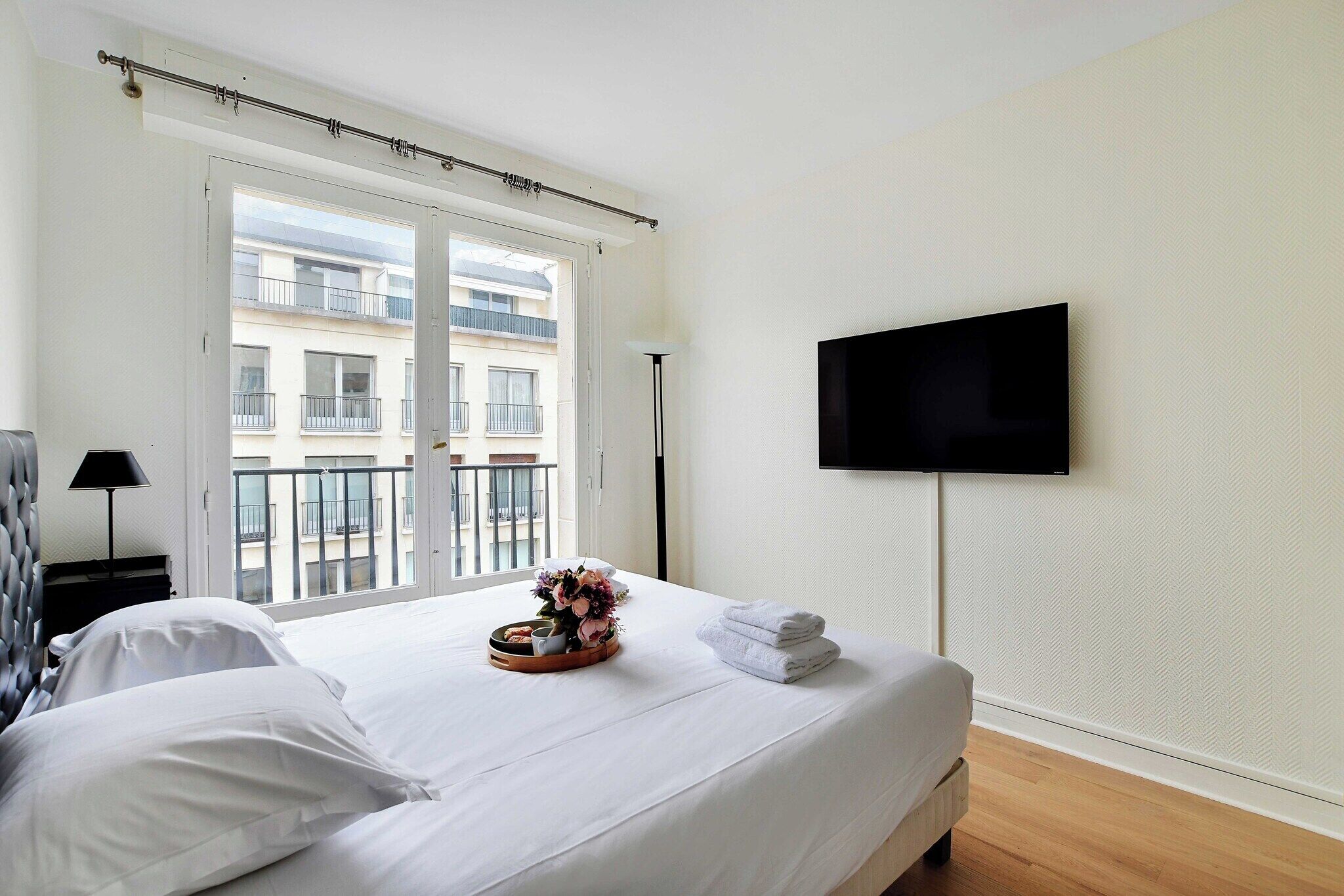 A hotel room with a bed, a flat-screen TV mounted on the wall, a window with a view of a balcony, and a bedside table with a lamp and a tray with towels and flowers.