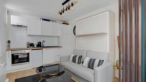 A modern kitchen with white cabinets, a built-in oven, and a coffee machine. There is a grey sofa with striped pillows and a black coffee table. - Cosy Apt - 2P - République / Marais (Paris)