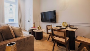 Una sala de estar moderna con un sofá, una mesa de comedor para dos personas, un televisor montado en la pared y una ventana con cortinas.
