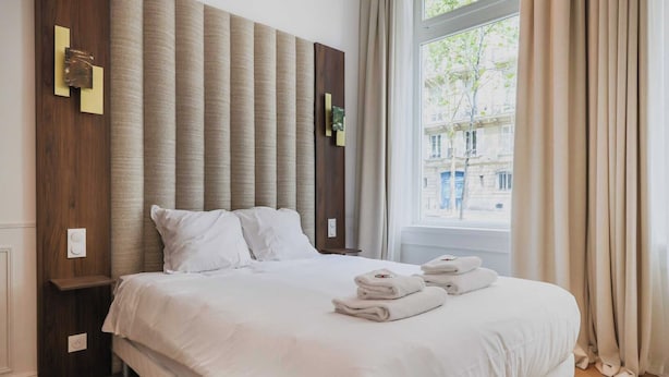 Una cama bien hecha con sábanas blancas y toallas dobladas, cabecera de madera y ventana con cortinas.