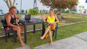 Dos personas sentadas en una mesa al aire libre con una computadora portátil, en un área con césped y un edificio blanco al fondo.