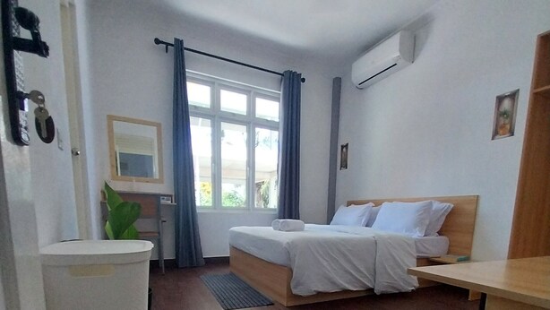 Habitación de hotel con cama, escritorio, cubo de basura, ventilador de techo y ventana con cortinas.