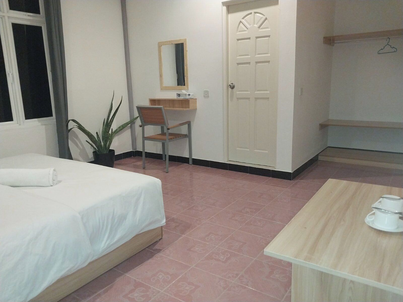 A hotel room with a bed, a desk, a chair, a potted plant, a wardrobe, and a small table with a cup and saucer.