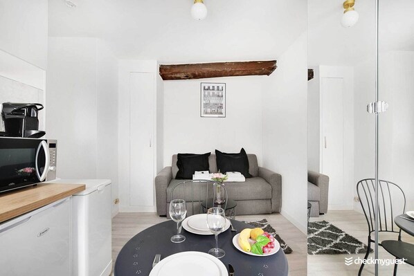 Una cocina moderna con microondas, tostadora y cafetera. Hay un comedor con una mesa dispuesta para dos personas, dos copas de vino y un plato de fruta. Un sofá gris y una silla negra se observan al fondo.