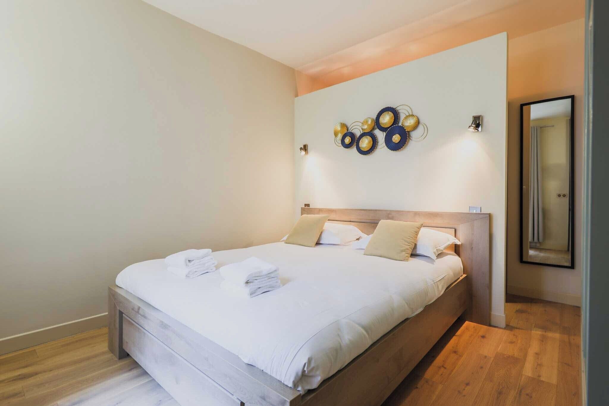 Una cama bien hecha con sábanas blancas y cabecera de madera en una habitación moderna con pared de color claro y una obra de arte decorativa sobre la cama.