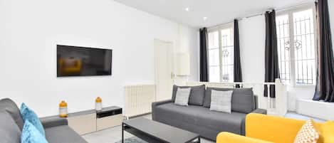 Una sala de estar moderna con un sofá gris, un sillón amarillo, una mesa de centro y un televisor montado en la pared.