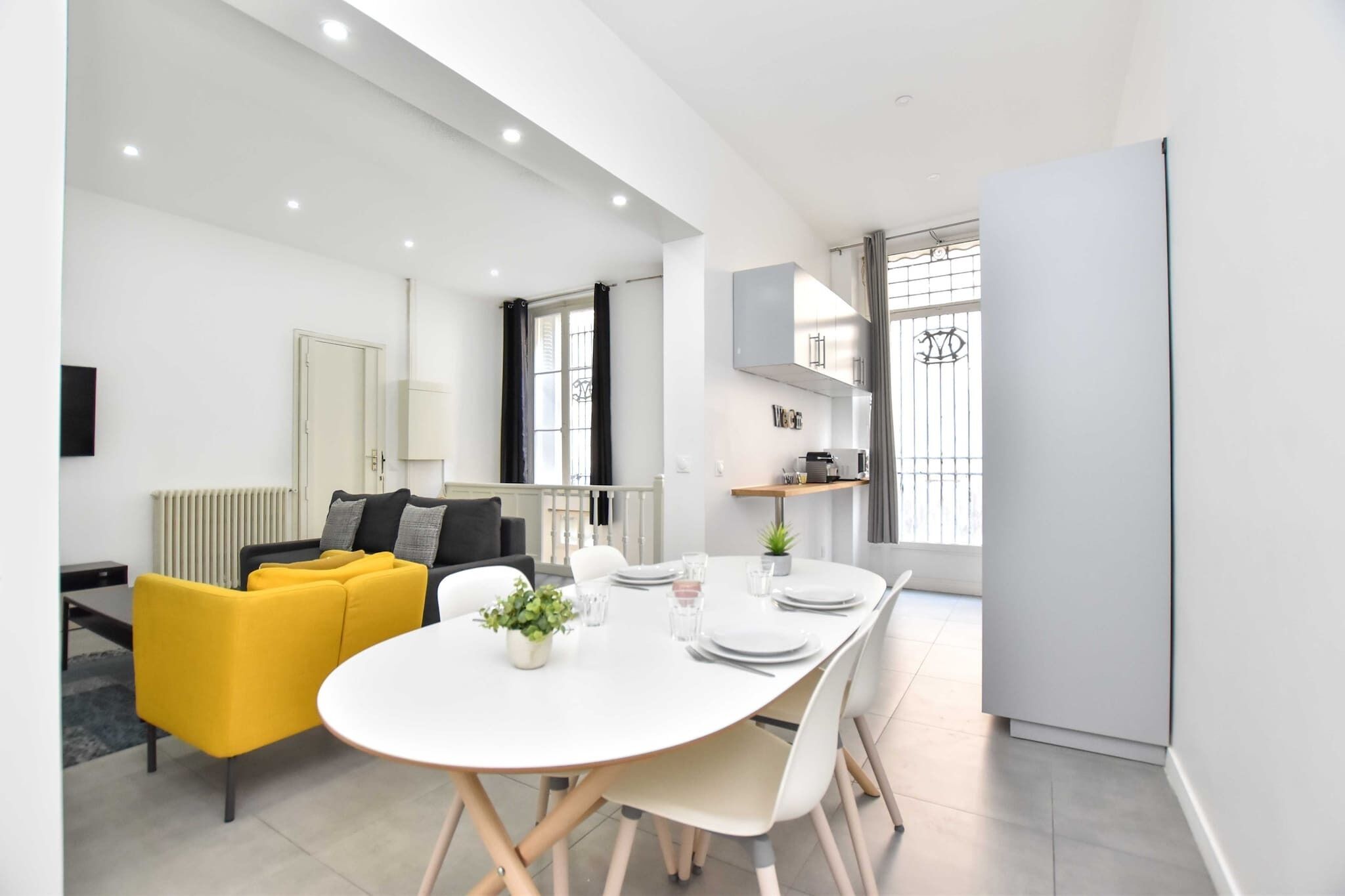 A modern, well-lit living room with a yellow sofa, a dining table set for four, and a kitchen area with a refrigerator and cabinets.