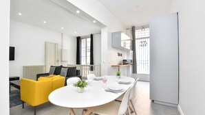 Un salon moderne et bien éclairé, avec un canapé jaune, une table à manger dressée pour quatre personnes et un coin cuisine équipé d’un réfrigérateur et de placards.
