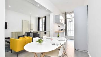 Un salon moderne et bien éclairé, avec un canapé jaune, une table à manger dressée pour quatre personnes et un coin cuisine équipé d’un réfrigérateur et de placards.