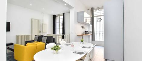A modern, well-lit living room with a yellow sofa, a dining table set for four, and a kitchen area with a refrigerator and cabinets.