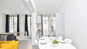 Un espace repas moderne avec une table blanche dressée pour quatre personnes, un fauteuil jaune et une vue sur la cuisine et le balcon.