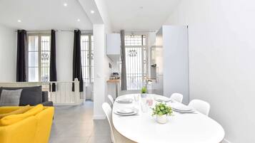 Un espace repas moderne avec une table blanche dressée pour quatre personnes, un fauteuil jaune et une vue sur la cuisine et le balcon.