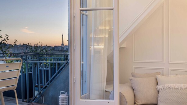 Een balkon met uitzicht op de stadshemel, een witte deur en een beige bank met kussens.