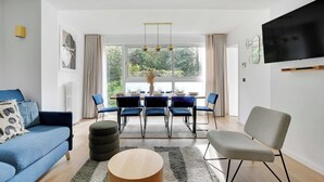 Un saló modern amb un sofà blau, un butac armchair gris, una taula de dinar amb caires i un televisor de pantalla plana fixat a la paret.