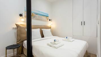 Un lit bien fait, avec du linge de lit et des serviettes blanches, une petite table de chevet, une lampe fixée au mur et un tableau représentant une scène de plage.