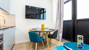 A modern dining area with a round table, two chairs, a flat-screen TV mounted on the wall, and a kitchen area with a sink and cabinets. - Intimate apartment - 1BR/4P - Oberkampf (Paris)