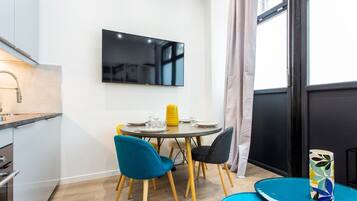 Un espace repas moderne comprenant une table ronde, deux chaises, un téléviseur à écran plat fixé au mur, ainsi qu’un coin cuisine avec un évier et des placards.