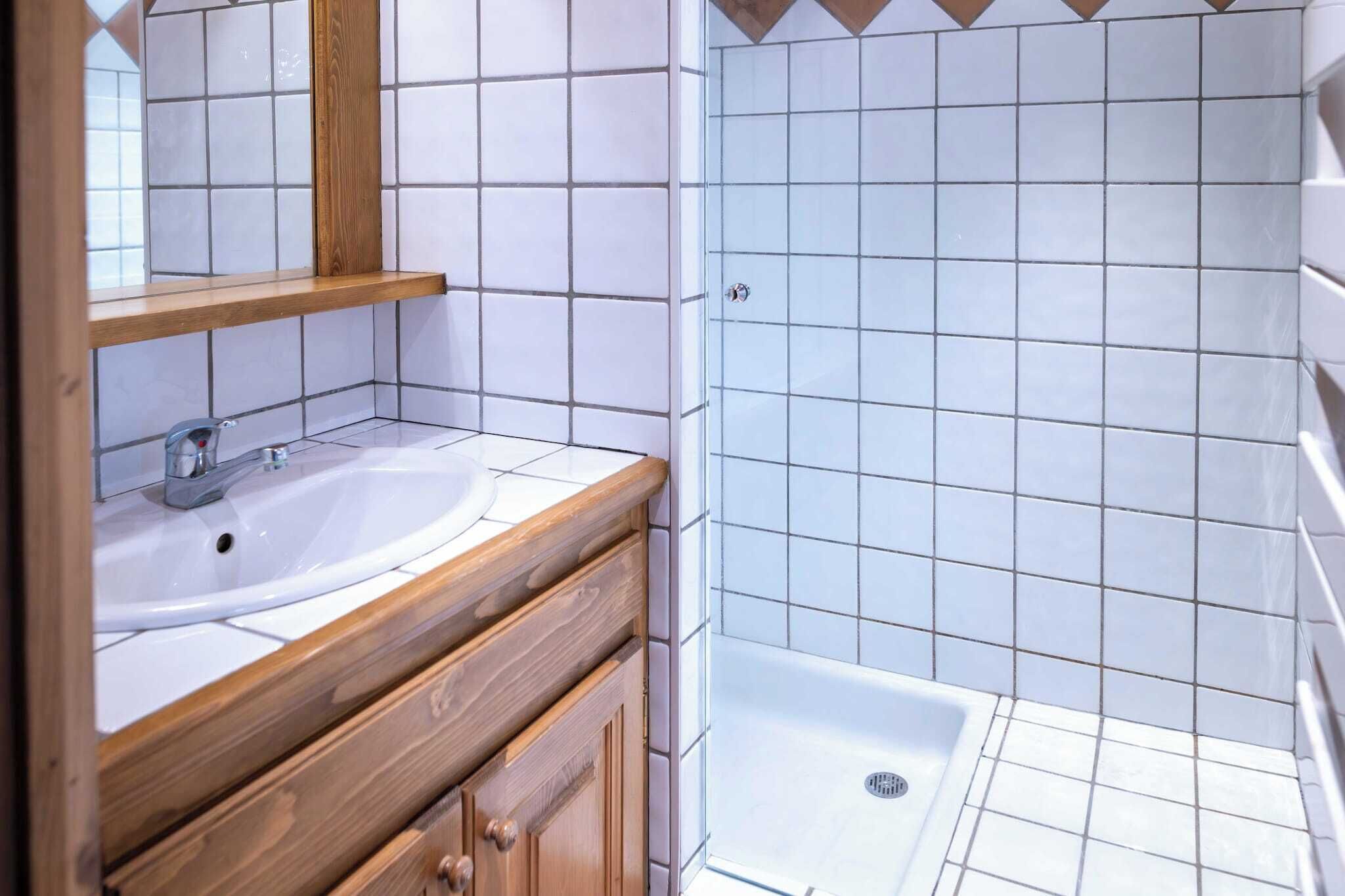 Un baño con lavamanos blanco, tocador de madera y zona de ducha con azulejos blancos.