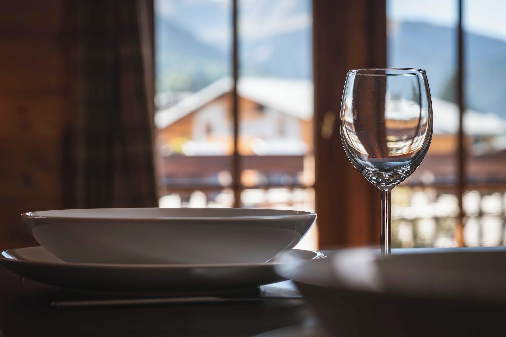 Una mesa de comedor con un tazón blanco y una copa de vino, frente a un interior de madera y vistas de edificios nevados.