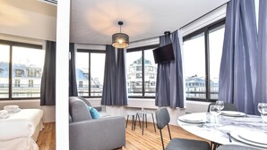 Ein modernes Hotelzimmer mit großem Fenster, einer Sitzecke, einem Esstisch für zwei Personen und einem an der Wand befestigten Flachbildfernseher.