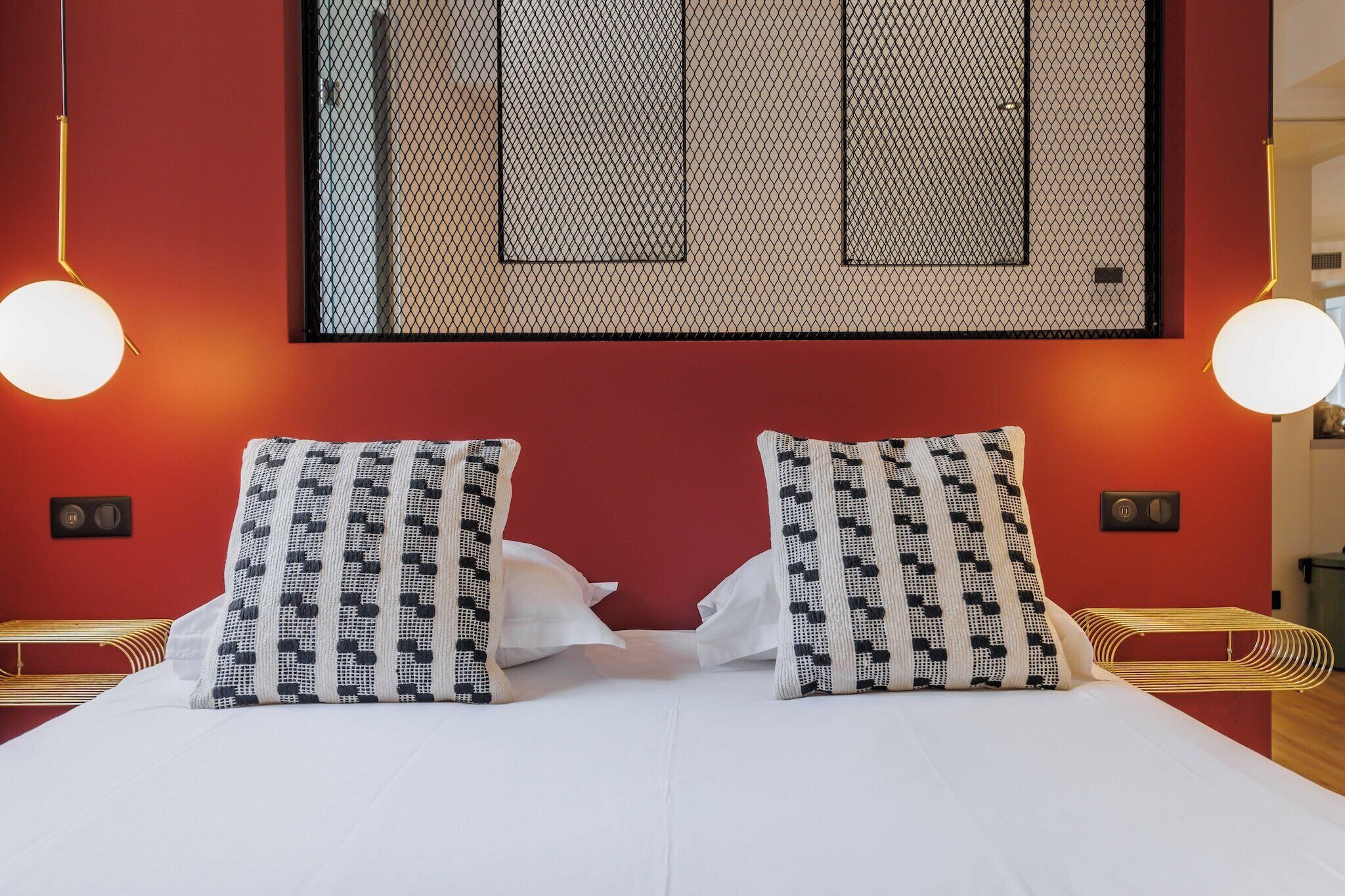 A bed with white bedding and patterned pillows, a red accent wall, and two pendant lights.