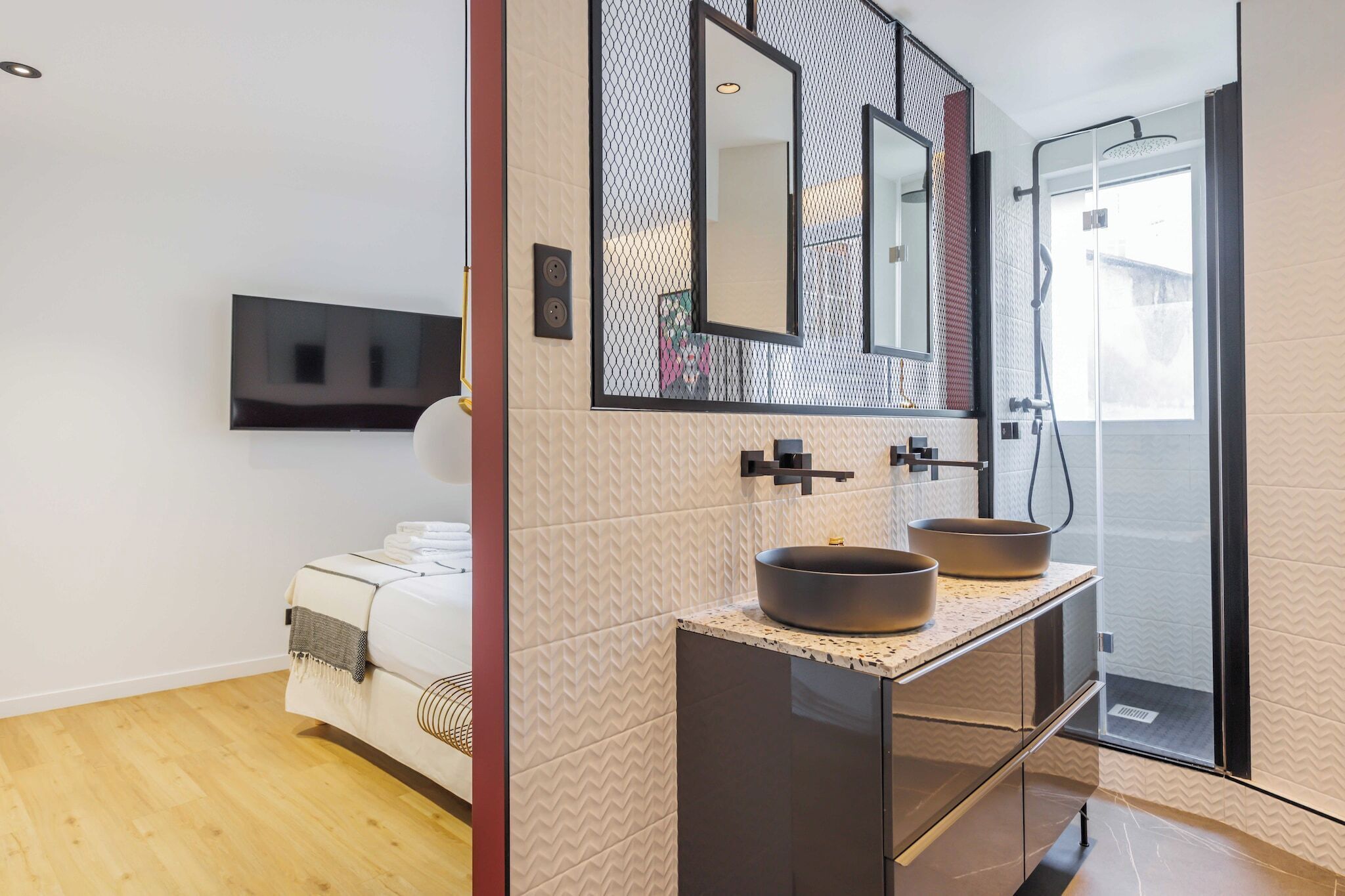 A modern bathroom with a large mirror, two vessel sinks, a shower, and a bed in the adjacent room.