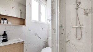 A bathroom with marble walls, a white toilet, a shower with a handheld showerhead, and a wooden shelf with toiletries. - Cosy apartment - 1BDR-4P - Ivry-sur-Seine (Ivry-sur-Seine)