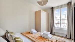 A hotel room with a bed, a wardrobe, a window with curtains, and a view of buildings outside. - Cosy apartment - 1BDR-4P - Ivry-sur-Seine (Ivry-sur-Seine)