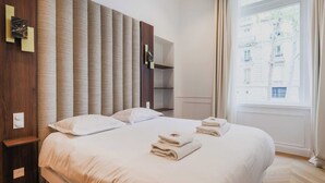 Habitación de hotel con una cama grande, cabecera de madera, mesita de noche y ventana con vistas a edificios.