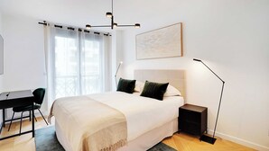 Een slaapkamer met een bed, nachtkastje, bureau, stoel en een groot raam met gordijnen.