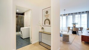 Een modern appartement met een glazen douche, een wasmachine, een eethoek met een tafel en stoelen, en een kunstwerk in een lijst aan de muur.