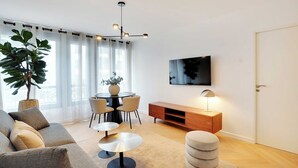 Een moderne woonkamer met een bank, een rond bijzettafeltje, een eettafel met stoelen, een potplant, een televisie aan de muur en een houten kast.