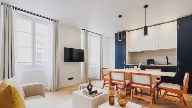 A modern living room with a sofa, coffee table, and dining area with wooden chairs and a table. There is a kitchen with dark cabinets and a flat-screen TV mounted on the wall.