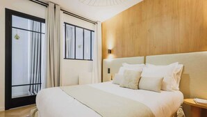 A modern hotel room with a large bed, a wooden headboard, a bedside table with a lamp, and a view of the city through large windows.