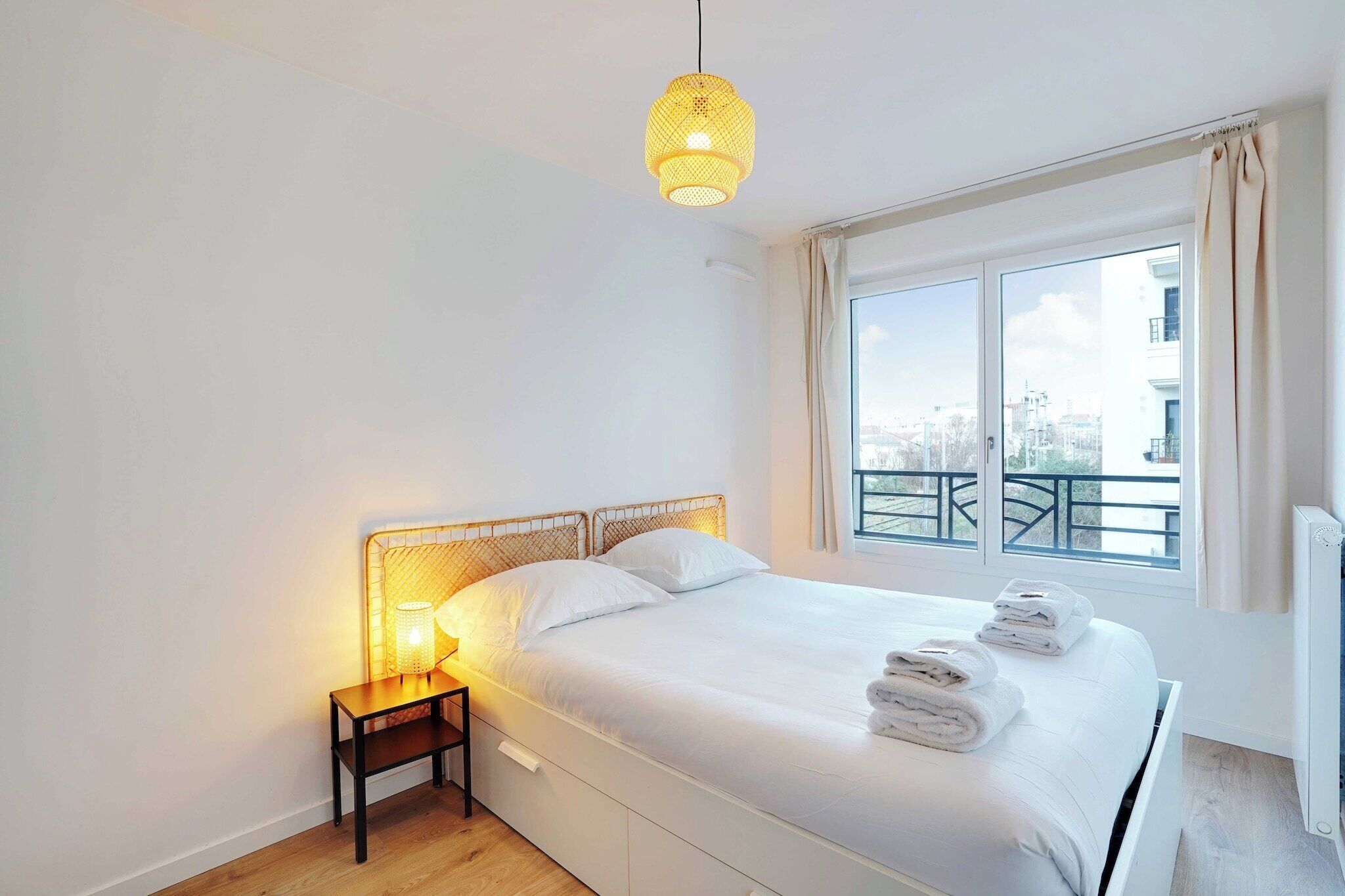 A neatly made bed with white linens and towels, a bedside table with a lamp, and a window with a view of buildings.