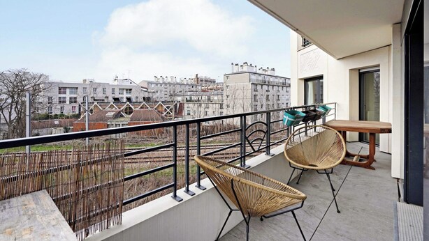 Ein Balkon mit Korbstühlen und einem Tisch, der auf Gleise und Wohnhäuser blickt.