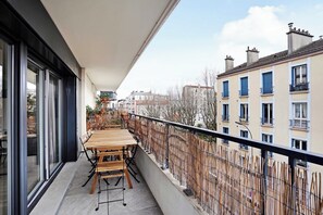 Un balcon avec une table et des chaises en bois, une porte vitrée et une vue sur les bâtiments voisins.