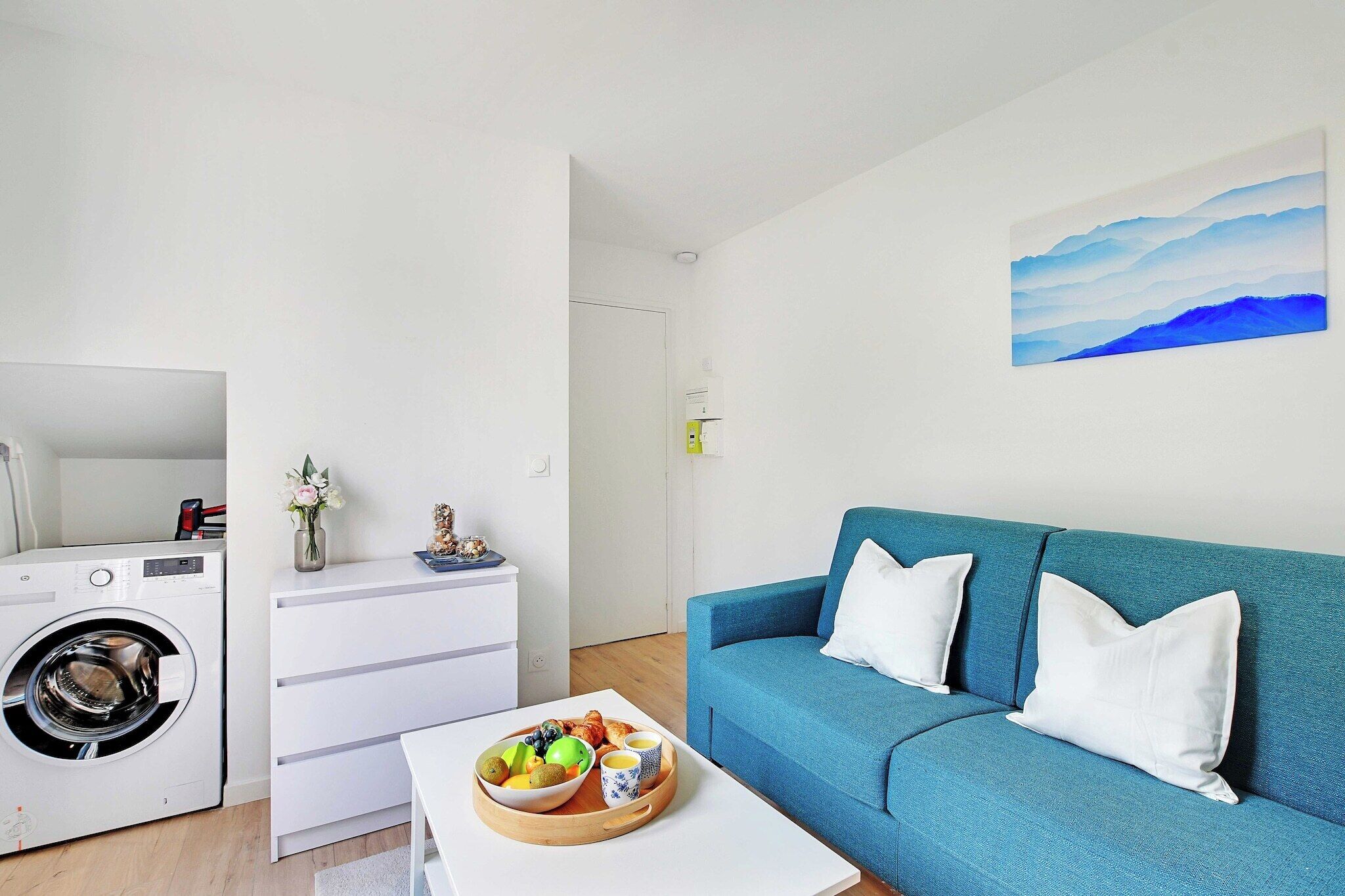 Un salon moderne avec un canapé bleu, un lave-linge, une table basse sur laquelle se trouvent des fruits et des tasses, et un tableau accroché au mur.