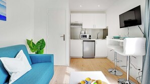 Un espacio habitable compacto con kitchenette, un sofá azul con un cojín blanco, una pequeña mesa de comedor con una bandeja de fruta y un televisor montado en la pared.