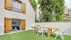 Un edificio blanco con contraventanas de madera, un balcón con césped artificial y una mesa de madera con sillas.