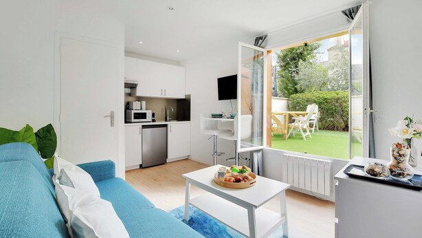 Una sala de estar moderna con un sofá azul, una mesa de centro blanca con un frutero, y vistas a un jardín a través de un ventanal.