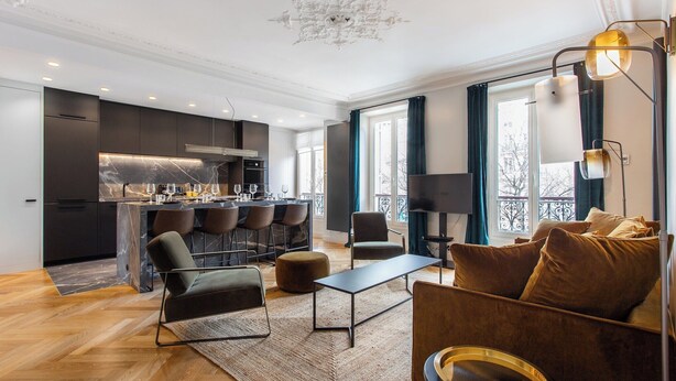 Ein modernes Wohnzimmer mit einer Couch, einem Couchtisch und einem Essbereich mit Stühlen, im Hintergrund eine Küche.