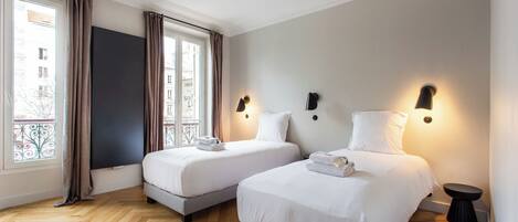 A hotel room with two beds, a large window with curtains, and a view of buildings outside.