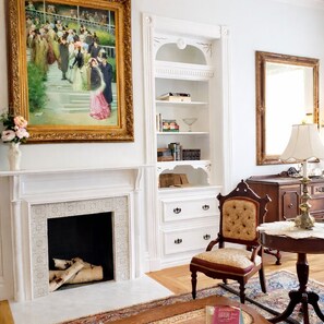A room with a fireplace, a bookshelf, a chair, a table, and a mirror. - The Queen On Main (Winters)